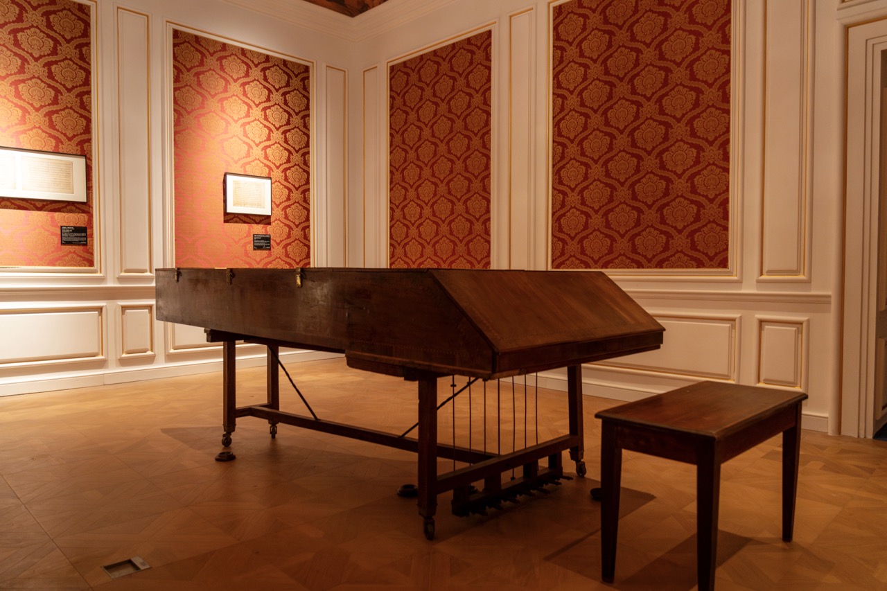 Rare early wooden harpsichord exhibited in room with red floral patterned wallpaper and wooden moldings.