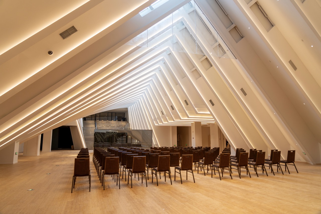Bright modern performance hall with layered sloped white ceiling linear lighting and neat rows of wooden audience chairs.