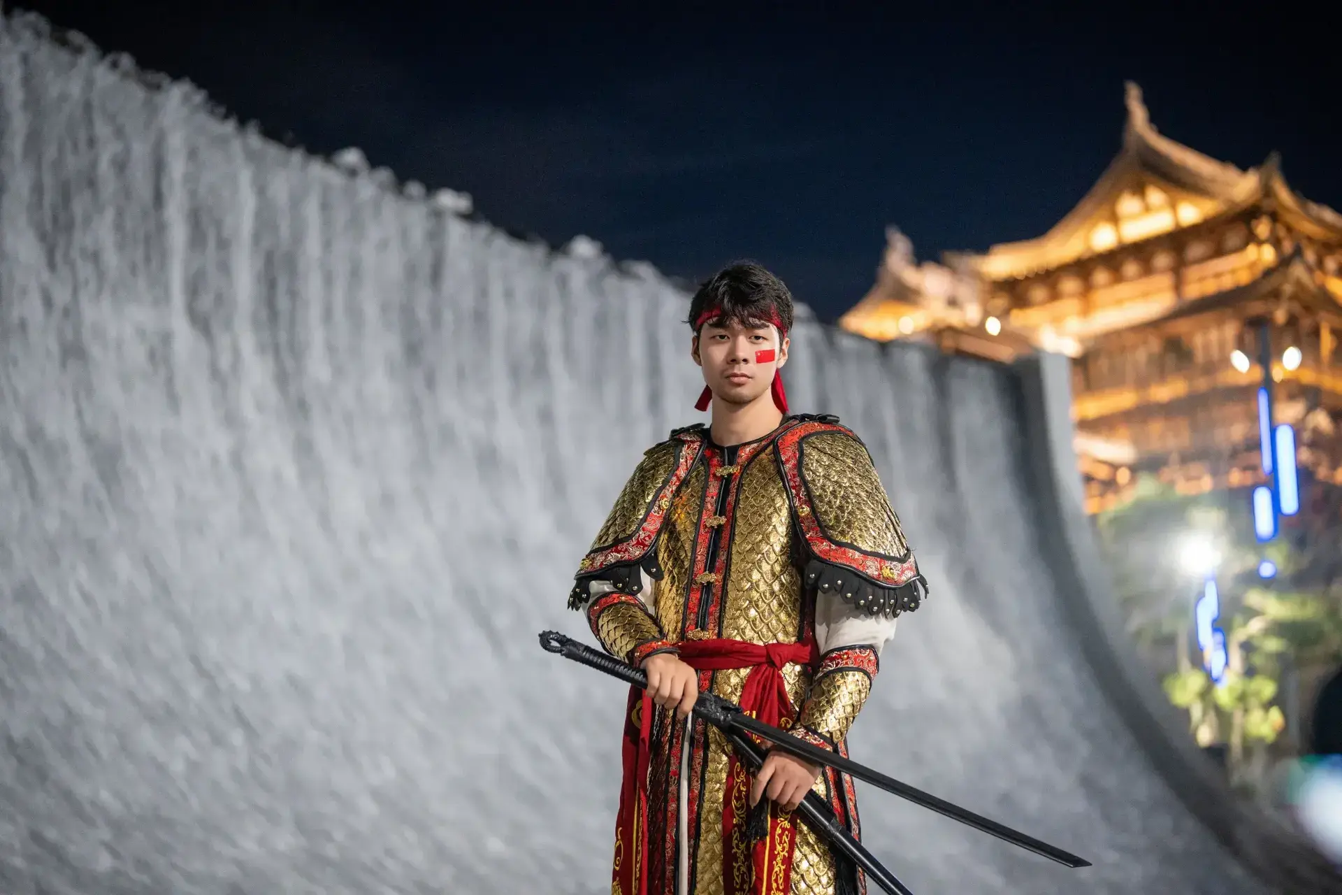 Young man in traditional golden scale armor holding swords, posing before giant waterfall wall with illuminated ancient wooden tower in Yanguan at night.