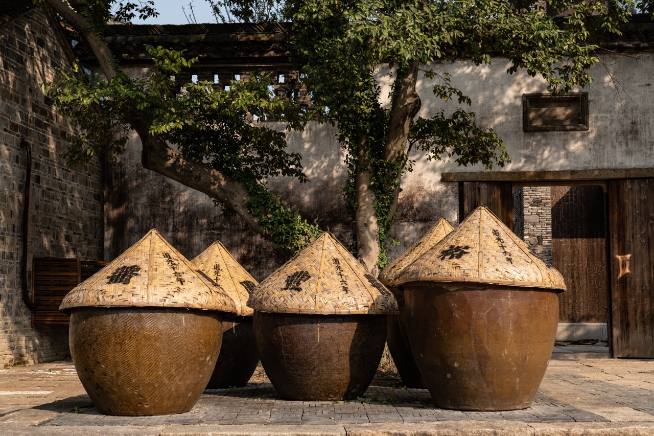 Row of earthen sauce vats topped with woven bamboo straw lids under old courtyard trees.