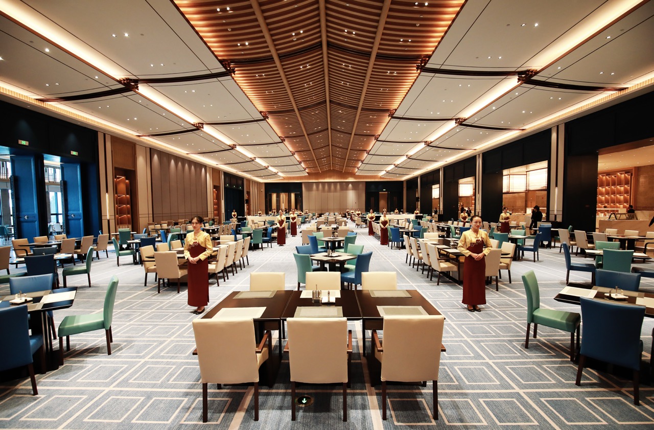 Bright modern buffet restaurant with layered wooden ceiling and multi-color upholstered chairs.